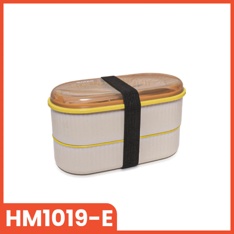 Kotak makan siang dua tingkat berwarna krem dengan aksen kuning di bagian tengah, tutup transparan berwarna cokelat muda, dan sendok garpu yang diletakkan di dalamnya, diikat dengan tali pengunci elastis berwarna hitam.