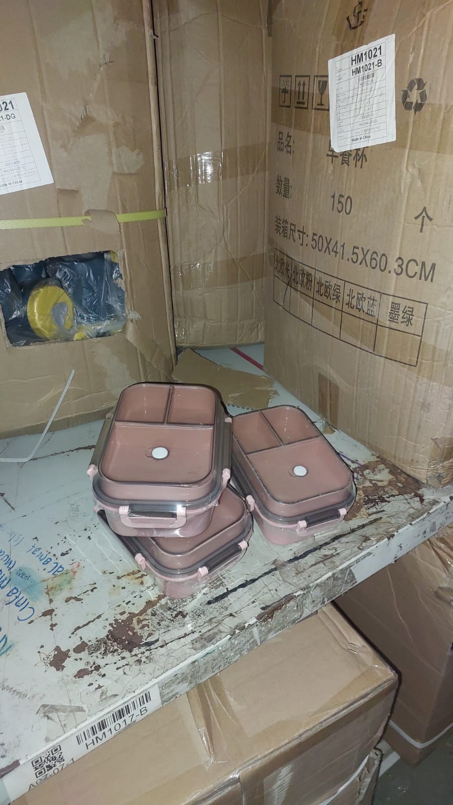 Stack of pink food containers on a cluttered surface with cardboard boxes and labels in the background.