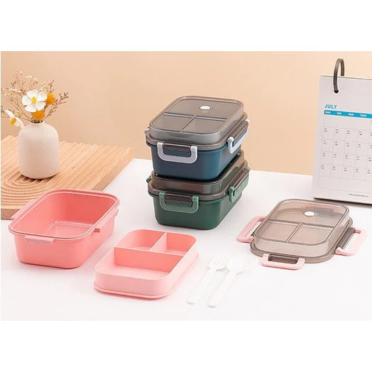 Dua set kotak makan siang dua tingkat di meja, satu set berwarna pink yang dibongkar dengan wadah, baki sekat, dan sendok garpu putih di samping. Set lainnya adalah dua kotak makan siang yang ditumpuk, satu berwarna teal dan satu lagi berwarna hijau tua, keduanya dengan tutup transparan dan tali pengunci. Sebuah vas bunga kecil dan kalender bulan Juli terlihat di latar belakang.