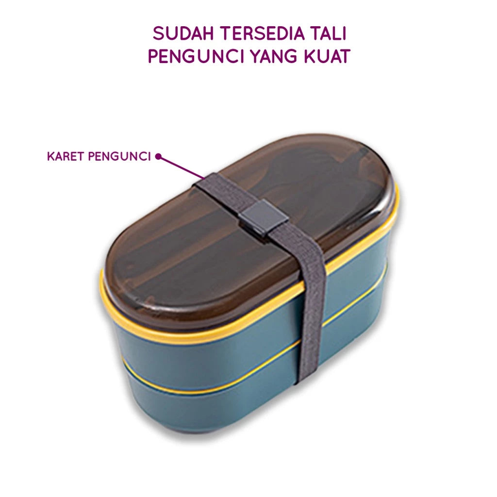 Kotak makan siang dua tingkat berwarna teal dengan aksen kuning di bagian bawah, tutup transparan, dan sendok garpu yang diletakkan di dalamnya, diikat dengan tali pengunci elastis berwarna abu-abu gelap.