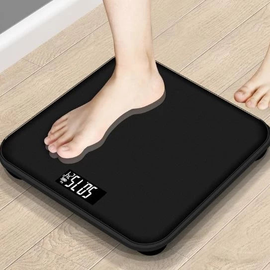 Timbangan tampak modern dengan desain minimalis dan sudut yang membulat. Latar belakang menunjukkan bagian sudut sebuah ruangan, kemungkinan besar kamar mandi atau kamar tidur.