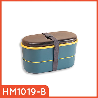 Kotak makan siang dua tingkat berwarna teal dengan aksen kuning di bagian bawah, tutup transparan, dan sendok garpu yang diletakkan di dalamnya, diikat dengan tali pengunci elastis berwarna abu-abu gelap.