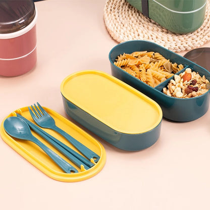 Kotak makan siang dua tingkat berwarna teal dan kuning dengan tutup kuning cerah, dibuka menampilkan bagian atas berisi pasta fusilli dan kacang-kacangan, dan sendok garpu teal terpisah di wadah kuning. Sebuah kotak makan siang lain berwarna hijau tua terlihat di latar belakang.