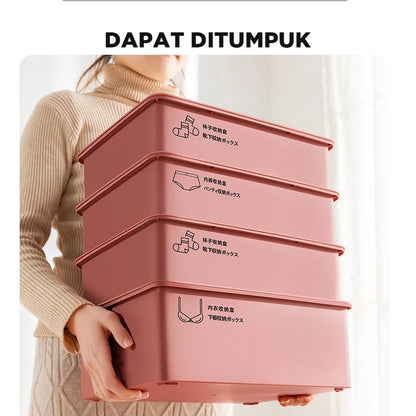  "Seorang wanita ber turtleneck coklat berdiri di depan tumpukan empat kotak organizer penyimpanan berwarna merah muda salmon yang dapat ditumpuk. Kotak-kotak tersebut memiliki simbol dan teks dalam bahasa Mandarin/Jepang yang menunjukkan fungsinya untuk menyimpan pakaian dalam, bra, atau kaus kaki. Terdapat tulisan 'DAPAT DITUMPUK' berwarna hitam di bagian atas gambar."
