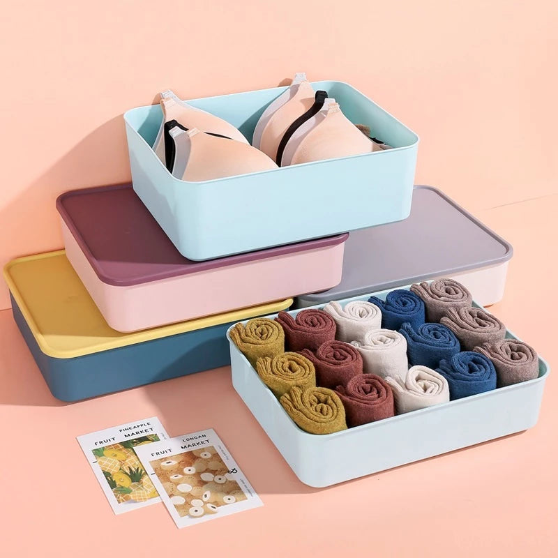 Set kotak organizer penyimpanan aneka warna di atas meja putih. Terlihat satu kotak biru muda diisi bra krem, ditumpuk di atas kotak ungu dan kuning. Di sampingnya, kotak biru muda lain berisi kaus kaki aneka warna digulung rapi, seperti merah marun, biru, krem, dan kuning mustard. Dua kartu bergambar buah-buahan tergeletak di depan kotak-kotak tersebut."