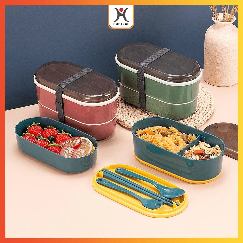 Dua kotak bekal Hoptech bertingkat dengan penutup transparan dan tali pengikat elastis; satu berwarna merah marun dan satu hijau tua. Di bagian depan, tampak kotak bekal terbuka berwarna teal dengan buah beri dan lemon, serta kotak bekal teal lainnya berisi pasta dan biji-bijian, lengkap dengan set sendok garpu biru di wadah kuning. Latar belakang berwarna hijau mint dan abu-abu