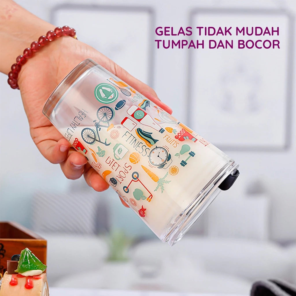 Sebuah tangan memegang gelas tumbler kaca transparan berisi cairan putih, dengan desain ilustrasi berwarna-warni bertema hidup sehat seperti sepeda, buah, sayur, barbel, dan tulisan "HEALTHY LIFE", "FITNESS", "DIET", "SPORT". Gelas menampilkan fitur anti-tumpah dan anti-bocor.