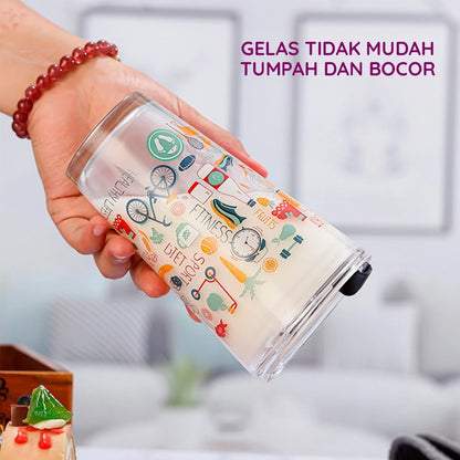 Sebuah tangan memegang gelas tumbler kaca transparan berisi cairan putih, dengan desain ilustrasi berwarna-warni bertema hidup sehat seperti sepeda, buah, sayur, barbel, dan tulisan "HEALTHY LIFE", "FITNESS", "DIET", "SPORT". Gelas menampilkan fitur anti-tumpah dan anti-bocor.