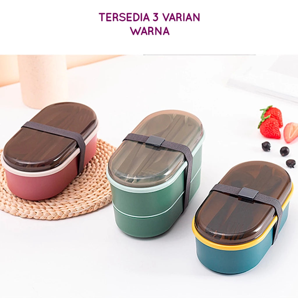  Tiga kotak makan siang dua tingkat dalam varian warna: merah muda gelap, hijau tua, dan teal-kuning, semuanya dengan tutup transparan dan tali pengikat elastis berwarna cokelat. Kotak makan siang diletakkan di atas meja dengan alas anyaman dan stroberi segar serta blueberry di latar belakang.